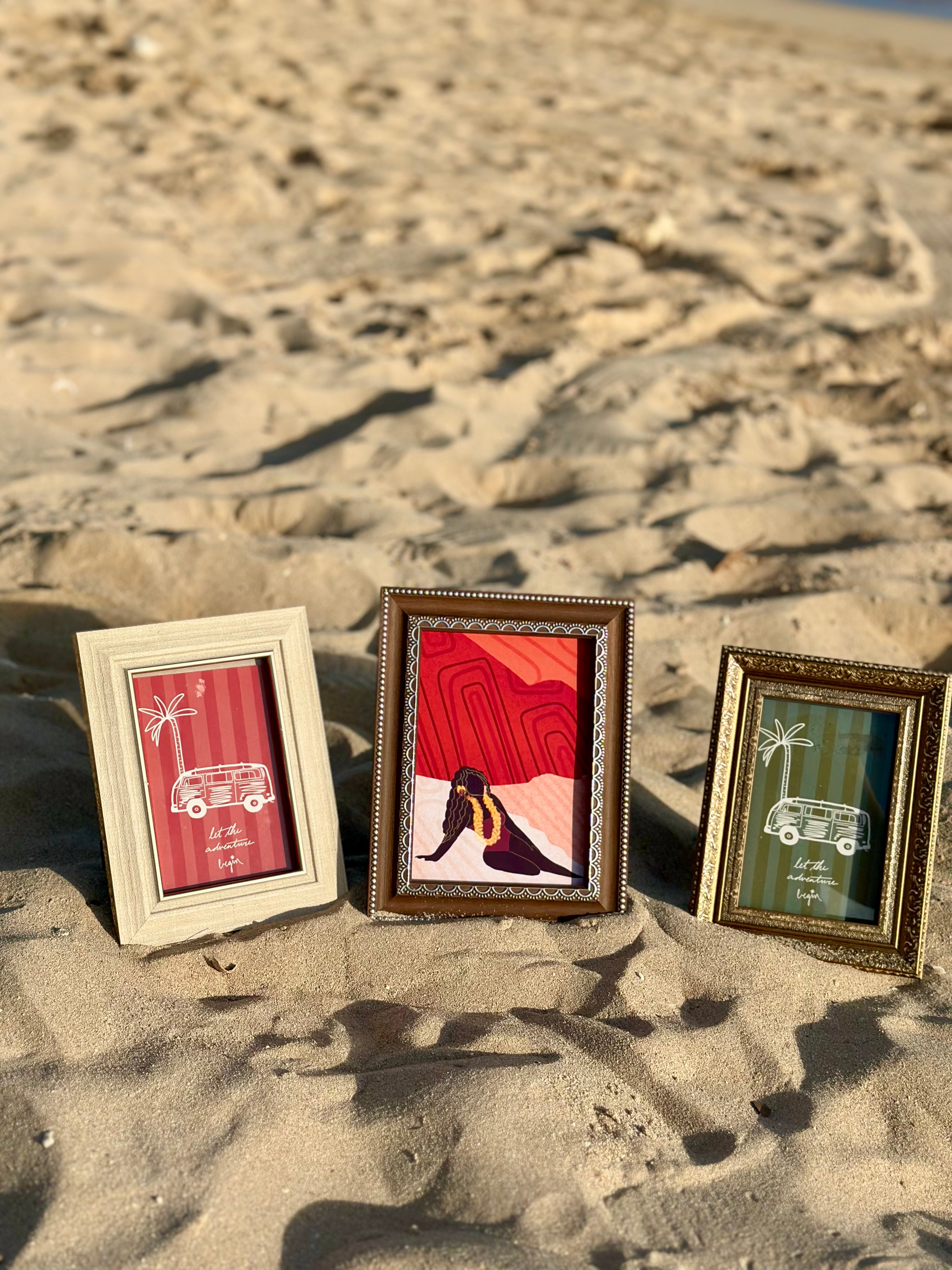 Three small decorative picture frames with vintage-style borders are arranged on sandy ground outdoors. The frames contain colorful art prints, including red, pink, green, and black-and-white designs.